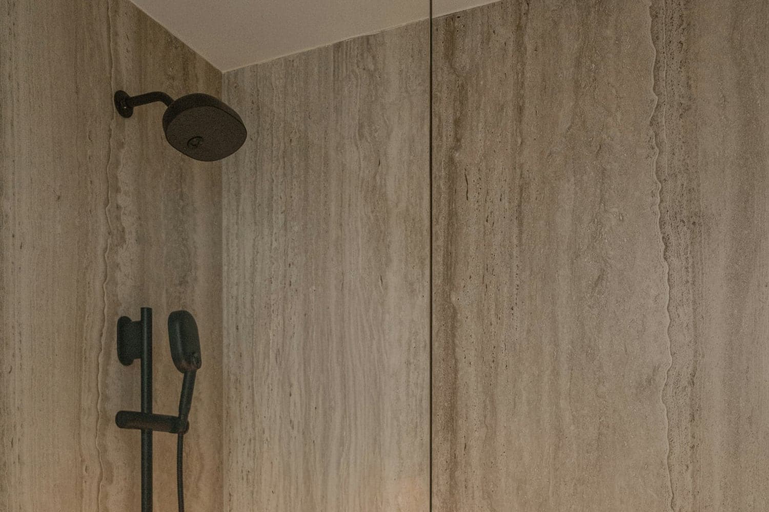 Détail de douche en travertin avec paroi vitrée et robinetterie noire.