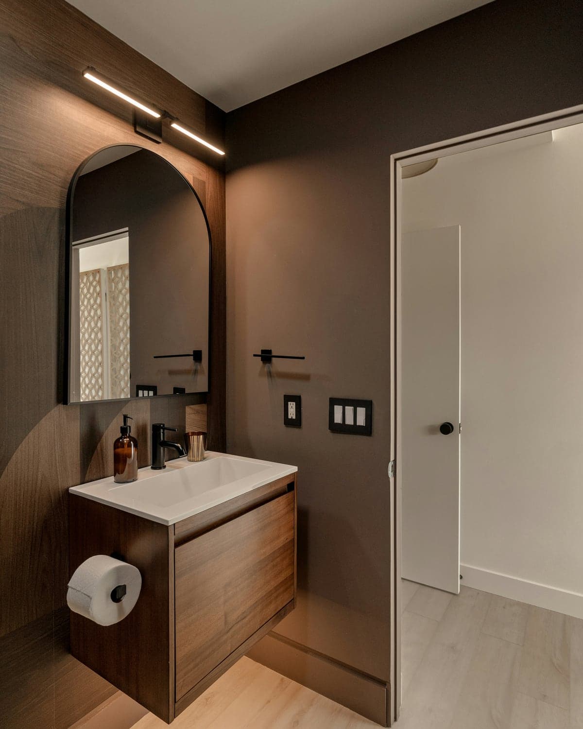 Salle de bain compacte avec meuble-lavabo en bois, miroir arqué et robinetterie noire.